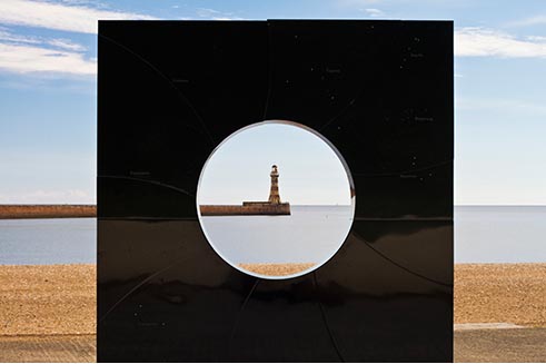 Photograph of Roker lighthouse cleverly framed by a circular hole in a modern sculpture. 

